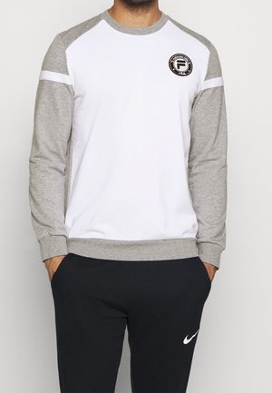 Mand iført langærmet hvid og grå sweatshirt med rund hals og patch-logo, kombineret med sorte Nike joggingbukser med hvid swoosh-logo.