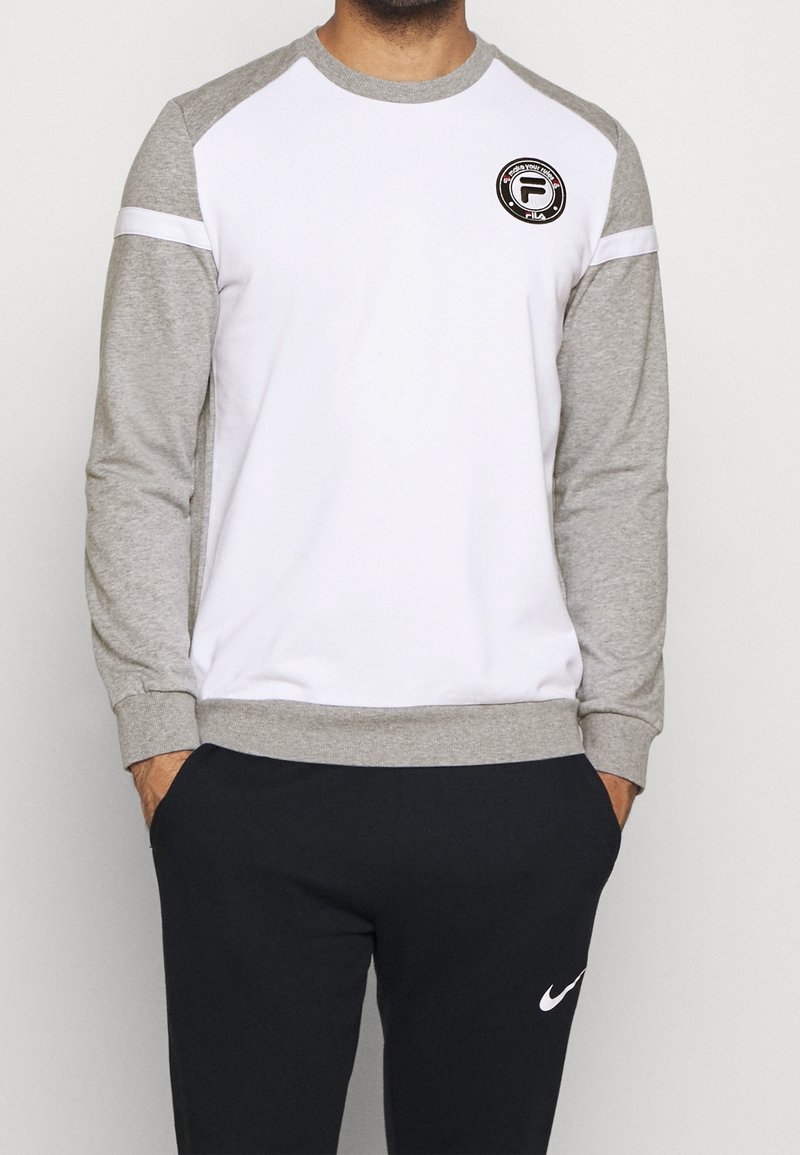 Homme portant un sweat-shirt à manches longues blanc et gris avec un col rond et un logo écusson, associé à un pantalon de survêtement Nike noir avec un logo swoosh blanc.