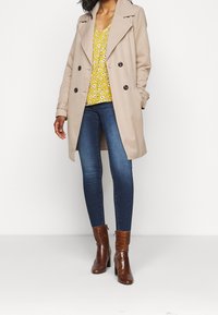 Beige trenchcoat med dubbelknäppt design, svarta knappar, bärs över en gul blommig topp, kombinerad med blå jeans och bruna ankelstövlar.