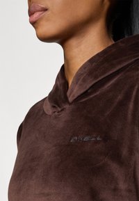 Brauner Kapuzenpullover aus weichem, plüschigem Material mit hohem Kragen und gesticktem Logo. Nahaufnahme des Kragens und der Stoffstruktur.
