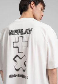 T-shirt bianca in cotone con grande testo grafico nero "REPLAY" e simboli sul retro. Vestibilità comoda con maniche corte e scollatura rotonda.