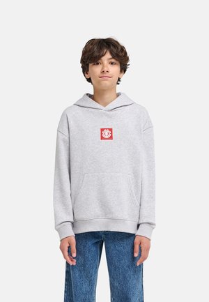 Młoda osoba o średniej długości brązowych włosach nosi jasnoszarą bluzę z czerwonym logo i niebieskie dżinsy, stojąc na jednolitym tle.
