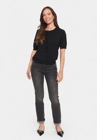 Svart kortärmad kofta med framknäppning, matchad med mörkgrå jeans med uppvikt nederkant och spetsiga svarta klackar. Slät textur genom hela plagget.
