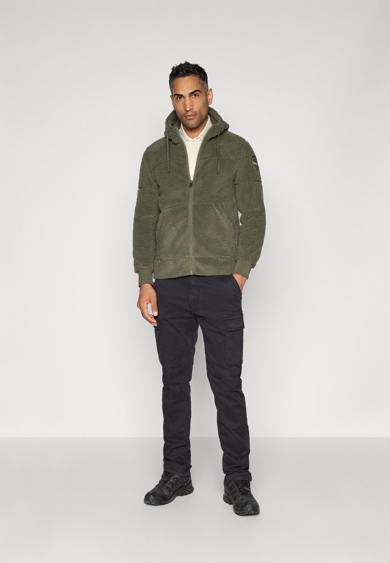 Mann trägt eine olivgrüne Fleecejacke mit Reißverschluss und Kapuze, schwarze Cargo-Hosen und schwarze Sportschuhe und steht vor einem schlichten hellgrauen Hintergrund.