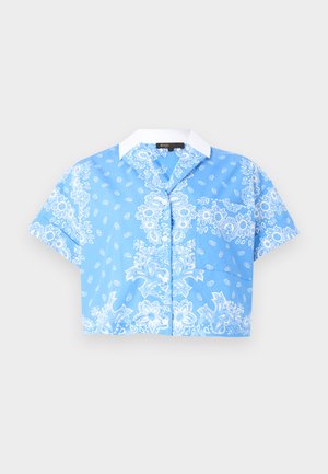 Kvinde stående iført en blå blomstret skjorte med korte ærmer, lyseblå vidde jeans med mønstre på siden, et brunt bælte og sorte stroppehæle.