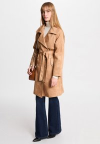 Manteau trench en faux suède beige avec de grands revers, ceinture à nouer et boutons argentés. Porté par-dessus un col roulé crème et associé à un jean évasé sombre.