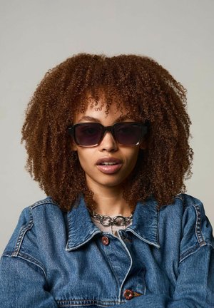 Gafas de sol negras de gran tamaño con lentes degradados y gruesos marcos de plástico. Se llevan con una chaqueta de denim y un collar de cadena de plata, contra un fondo neutro.