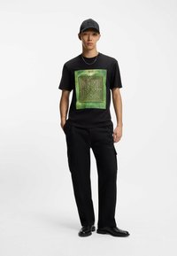 Zwarte t-shirt met een groen doolhofontwerp, korte mouwen, gecombineerd met zwarte cargo-broeken. Het model draagt een zwarte pet en zwarte schoenen.