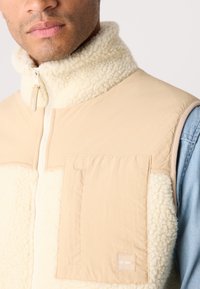 Beige fleece vest with high collar, featuring a textured cream panel, zip closure, and a large front pocket with a label.