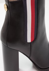 Bottine noire en cuir à talon haut avec panneau latéral élastique rouge, blanc et marine et détail de logo circulaire doré près du haut.
