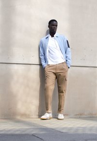 Helles blaues Jackett über einem weißen Hemd, beige Cargo-Hosen und weiße Sneakers. Glatte Stofftexturen mit einem Taschen-Detail an der Jacke.