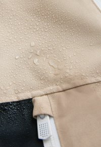 Beige water-repellent fabric with droplets; features a black section and a white zipper with a mesh accent. Smooth texture, matte finish.