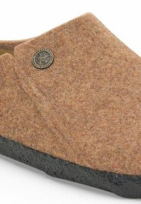 Pantofola di feltro marrone con superficie testurizzata, forma arrotondata, suola in gomma nera e un emblema metallico rotondo sul lato.