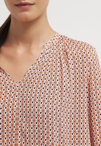 Blusa arancione con motivo geometrico, scollatura a V, con disegni bianchi e marroni scuri, dal tessuto morbido e dettagli arricciati sulle spalle.