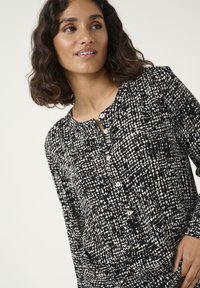 Černobílé vzorované blouse s kulatým výstřihem, zapínáním na knoflíky a nažehlenými detaily na ramenou. Měkká tkanina, dlouhé rukávy.