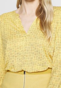 Gelbe Bluse mit einem Muster aus schwarzen und orangen geometrischen Linien; hat einen V-Ausschnitt und eine weite Passform. Weicher Stoff mit einer strukturierten Optik.