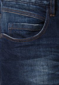 Mörk denimjeans med en tjock tygstruktur, som har en framficka, kontrastsömmar i orange och lätt blekning på ytan.