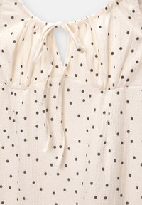 Blusa de color beige claro con lunares negros, de tejido texturizado, con cuello redondo y un cierre con lazo en la parte posterior. Presenta detalles fruncidos en los hombros.
