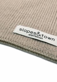 Tessuto lavorato a maglia beige con texture a costine, dotato di un'etichetta in tessuto bianco con testo nero che recita "slopes&town AMSTERDAM."