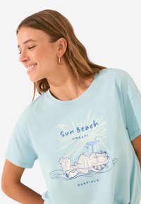T-shirt en coton bleu clair avec un graphisme d'un chat cartoon se prélassant près de l'eau, avec le texte "Sun Beach Amalfi" au-dessus et "Garfield" en dessous.