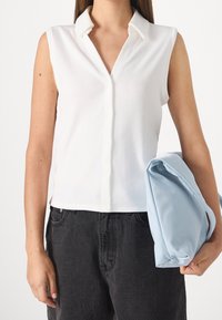 Femme portant un haut débardeur blanc à boutons et un jean noir, tenant un sac plié bleu clair sous son bras.