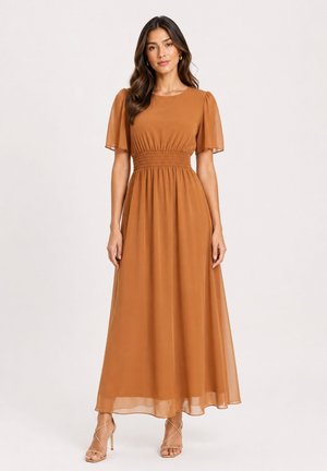 Donna che indossa un lungo vestito color ruggine con maniche corte svolazzanti e vita arricciata, in piedi con le braccia rilassate lungo i fianchi.