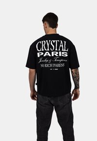 Černé oversized tričko s bílým textem na zadní straně: "CRYSTAL PARIS Šperky & Hodinky BEZ BOHATÝCH RODIČŮ". Krátké rukávy, volný střih.
