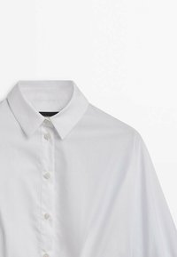 Chemise blanche à manches longues boutonnée, col pointu, quatre boutons visibles et texture de tissu lisse sur fond uni.