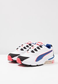 Chaussures de sport blanches avec des accents noirs, bleus et corail, en mesh et matériaux synthétiques, œillets en boucle noirs et amorti visible sous la semelle.