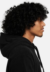 Schwarzer Fleece-Hoodie mit strukturiertem Material, ausgestattet mit einer Kordelzugkapuze und schwarzen Akzenten. Im Profil sind ausgeprägte Locken und ein glatter Teint zu sehen.