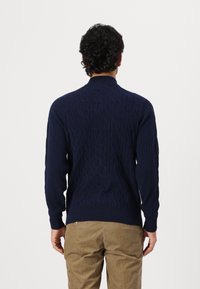 Granatowy sweter z dzianiny o fakturowanym wzorze w warkocze, z prążkowanym kołnierzem i mankietami, oraz dopasowanym kroju. Nosi się go na jasnych spodniach.