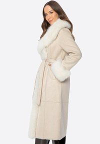 Beige lange jas met een witte faux fur kraag en manchetten, voorzien van een ceintuur in de taille en een gladde textuur.
