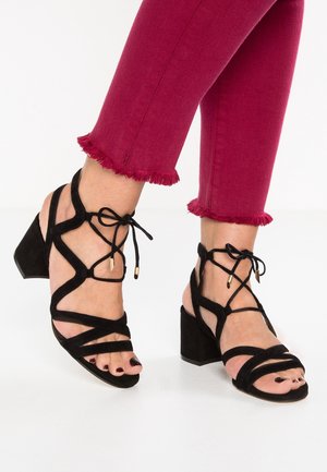 Ben iført røde cropped jeans med frynset kant og sorte remsandaler med blokhæl og ankelbind på hvid baggrund.