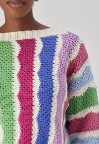 Strickpullover mit weitem Ausschnitt, der ein Zickzackmuster in den Farben Grün, Pink, Blau, Lila und Creme aufweist. Strukturierte Stoffdetails sind sichtbar.