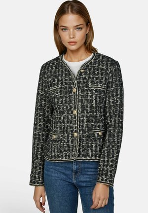 Jeune femme portant une veste boutonnée à motifs noir et blanc avec des boutons dorés et un jean bleu sur un fond uni.