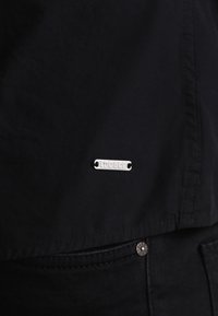 Zwart textiel met een gestructureerd oppervlak, voorzien van een zilveren metalen label met de tekst "EDC & CO" bevestigd nabij de zoom. Een subtiele, gepatternte detaillering is zichtbaar.