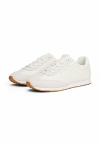 Un paio di sneakers basse color avorio con suole in gomma marrone chiaro e lacci bianchi, caratterizzate da un marchio discreto sul lato.