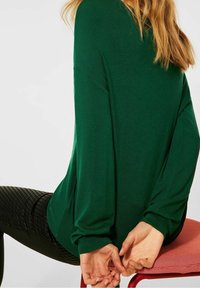 Grön stickad tröja med långa, lösa ärmar och avslappnad passform. Bärs med texturerade svarta leggings, sitter på en röd stol.