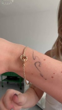 Bracciale in oro con un design attorcigliato e una chiusura circolare, indossato su un polso lentigginoso, accanto a un tatuaggio a forma di onda.