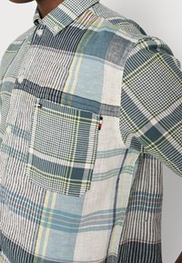 Camicia da uomo con bottoni in un patchwork di quadri blu, verdi e bianchi. Presenta una tasca sul petto e un colletto con un taglio casual.