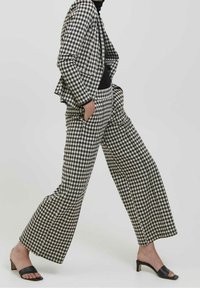 Ensemble blazer à carreaux noir et blanc et pantalon à jambes larges. Tissu texturé avec poches et coupe décontractée. Associé à des sandales à talons noires.