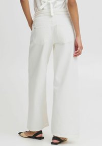 Pantalones de pierna ancha blancos con una textura de tela suave, que cuentan con dos bolsillos traseros y una cintura alta, combinados con zapatos slingback en negro y crema.