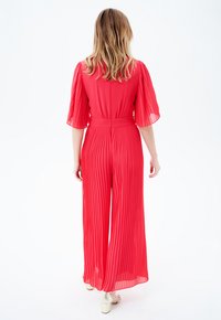 Mono rojo con pliegues, de mangas cortas y fluidas, cintura ceñida y diseño de pierna ancha. Textura suave con un tono vibrante.