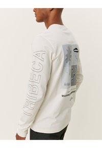 Kerman pitkähiha-t-paita, jossa on grafiikkasuunnittelu selässä ja "TRIBECA"-teksti vasemmassa hihassa. Pehmeä puuvillainen materiaali.