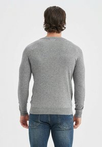 Pull en maille gris à manches longues, col rond, avec des poignets et une taille côtelés. Porté avec un jean en denim bleu, présentant une texture lisse.