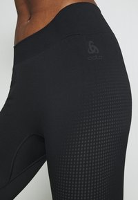 Schwarze Leggings mit glatter Textur, ausgestattet mit einem breiten Gummibund und einem dezenten Punktmuster an der Seite. Enthält ein unauffälliges Logo.