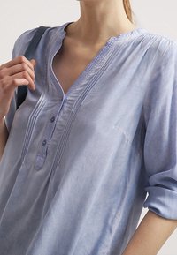 Kobieta w jasnoniebieskiej bluzce z długimi rękawami, zdobionej plisami i guzikami, trzyma ciemny pasek na swoim ramieniu.