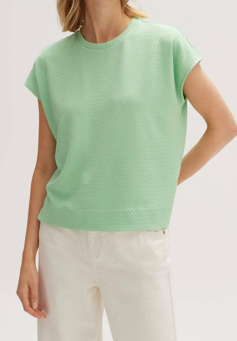Femme portant un haut texturé vert clair à manches courtes et un pantalon blanc taille haute, debout avec une main dans le dos.