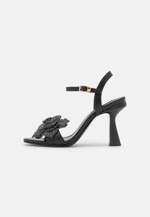 Sandalia de tacón alto negra con diseño floral, detalles de tachuelas doradas, correa ajustable en el tobillo y tacón ancho y resistente.
