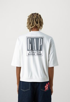 Persoon met blonde dreadlocks die een oversized wit GAP-shirt draagt met de tekst "ATHLETIC SINCE 1969" op de achterkant en donkere spijkerbroek.